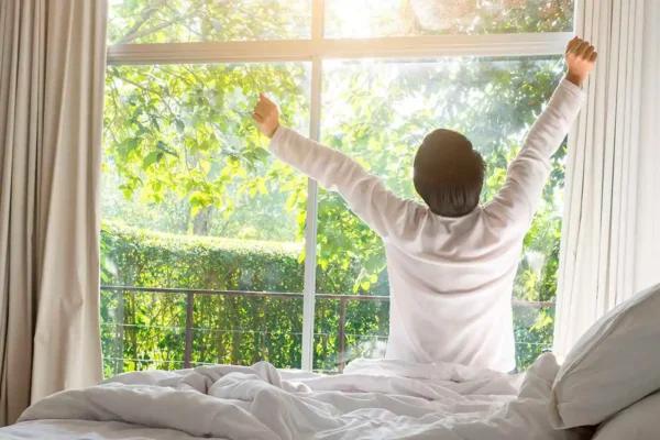 hombre que se despierta del sueño y se estira o despereza frente a una ventana con mucho sol, en un lugar tranquilo y mucha naturaleza, vestido de blanco y contento de haber dormido bien