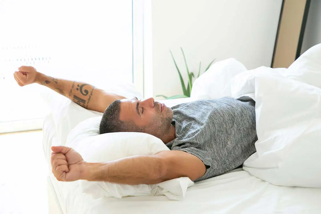 Hombre joven, despertando de su sueño, contento, plácido, con puños cerrados y levantando los brazos en su cama, dando la sensación de haber dormido bien, almohada y sabanas blancas y en una habitación muy iluminada.