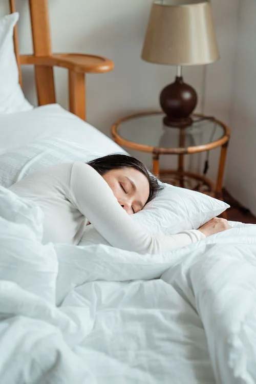 Mujer durmiendo placidamente, tapada con un edredón o acolchado mullido blanco, en pleno sueño con una remera larga blanca, en una habitación con paredes blancas y una lámpara con pantalla marron y una mesita de madera con un vidrio redondo.